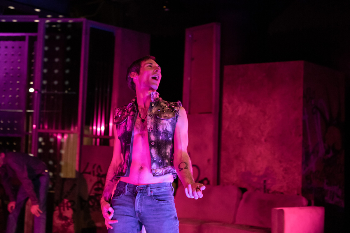 A man stands on stage in an acid wash vest and jeans as he speaks passionately. 