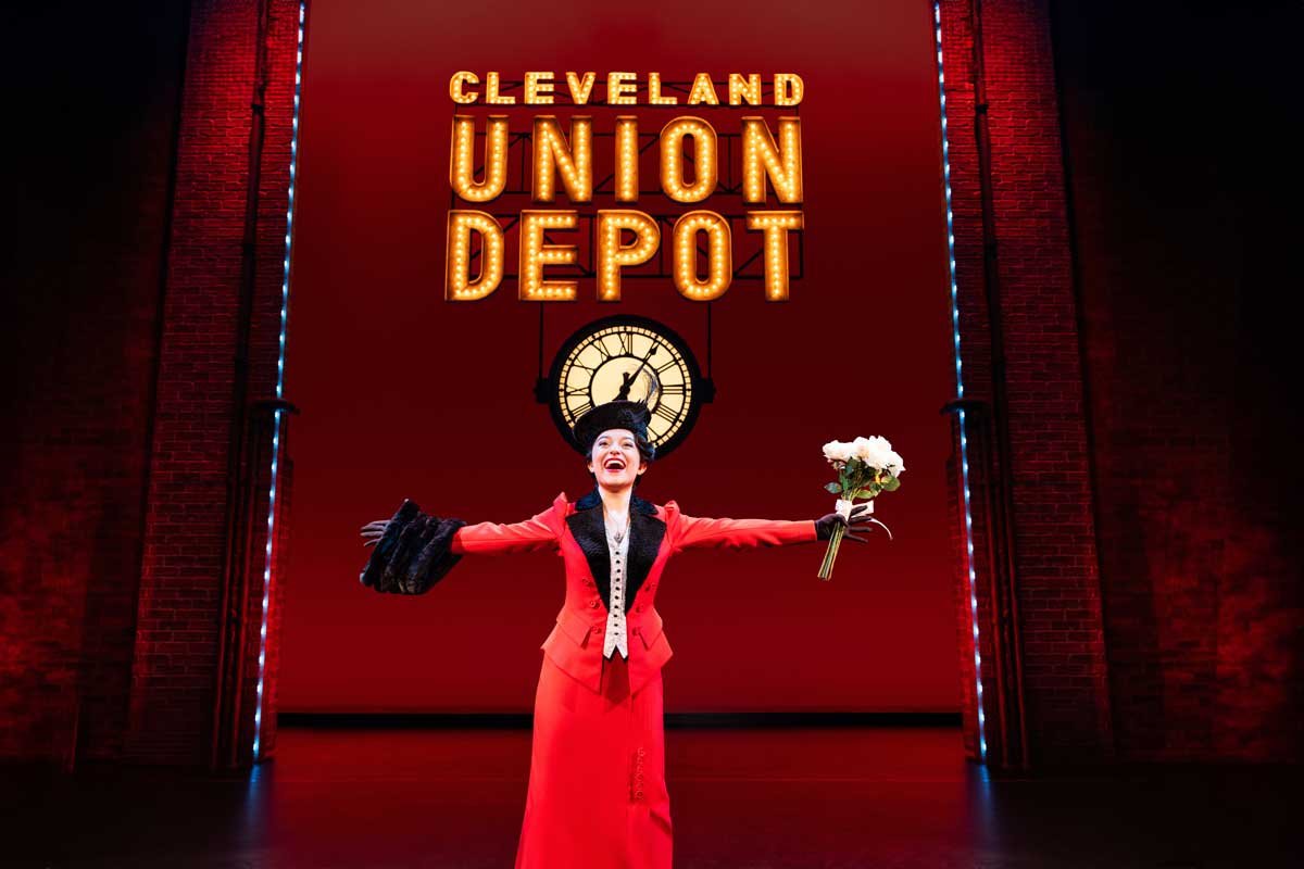 Fanny Brice wears a red dress holding her arms outstretched with a smile. She stands under a Cleveland Union Depot sign.