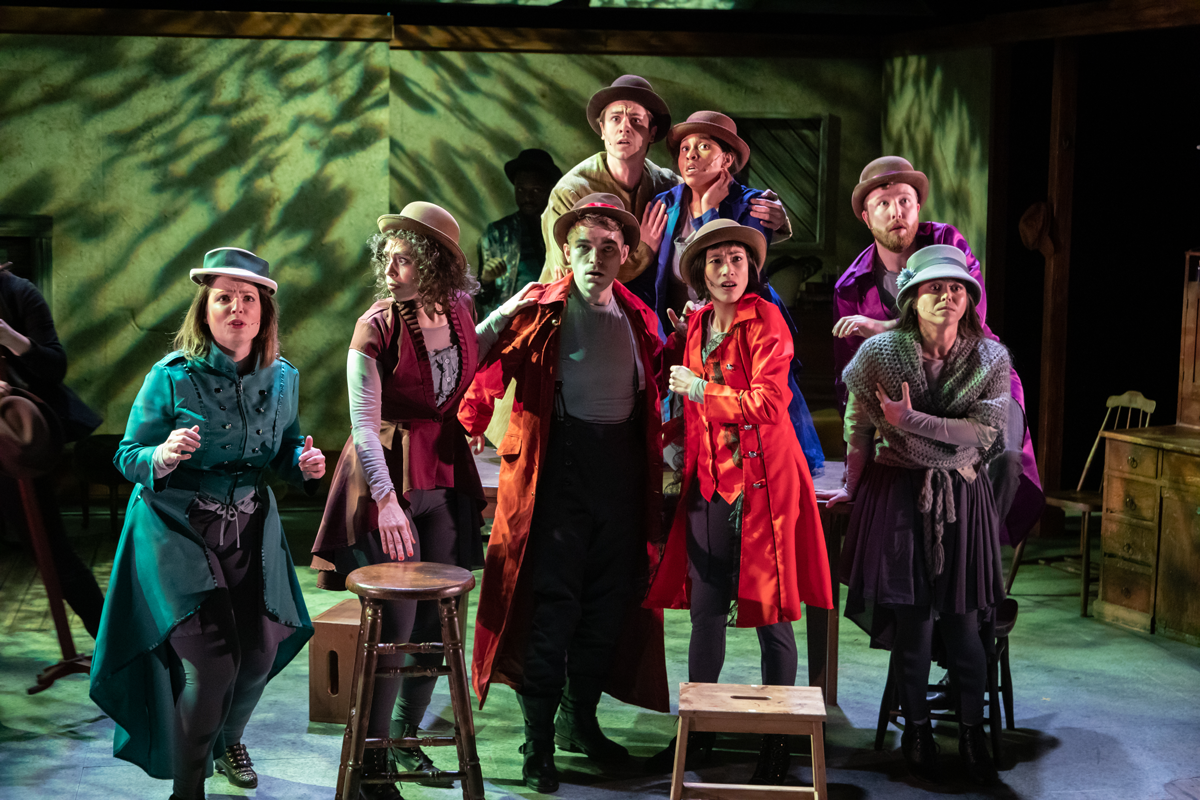 Eight actors on stage stand looking off into the distance with surprise with a forest shadow background.