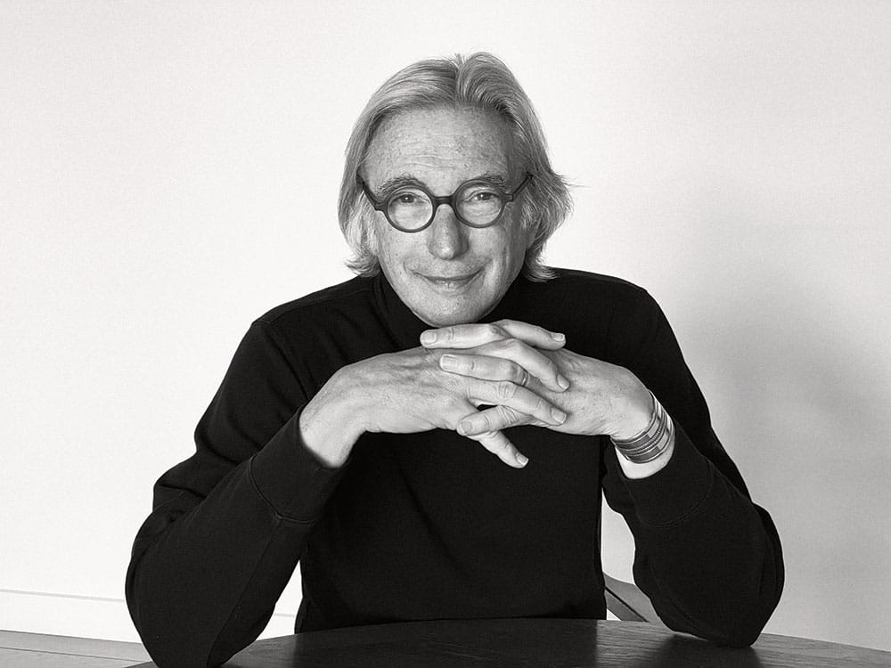 Michael Tilson Thomas in black and on a white background, hands clasped at a desk.