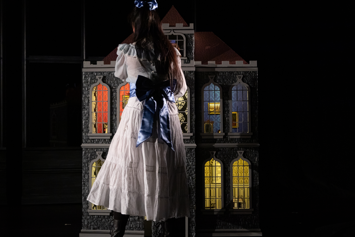 On a dark stage a young girl dressed in a 1910s dress stands in front of a doll house.