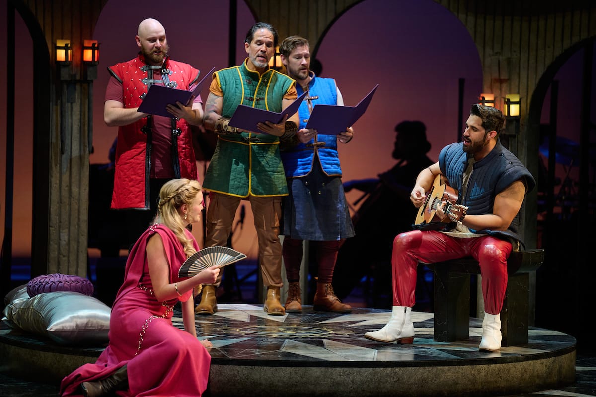 A group of men in bright middle age clothing sing and play guitar for a blonde woman in a pink dress, holding a fan.