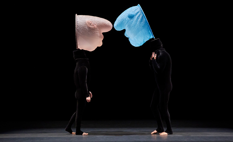 A black stage, two actors wearing black clothes face each other with large masks on their heads