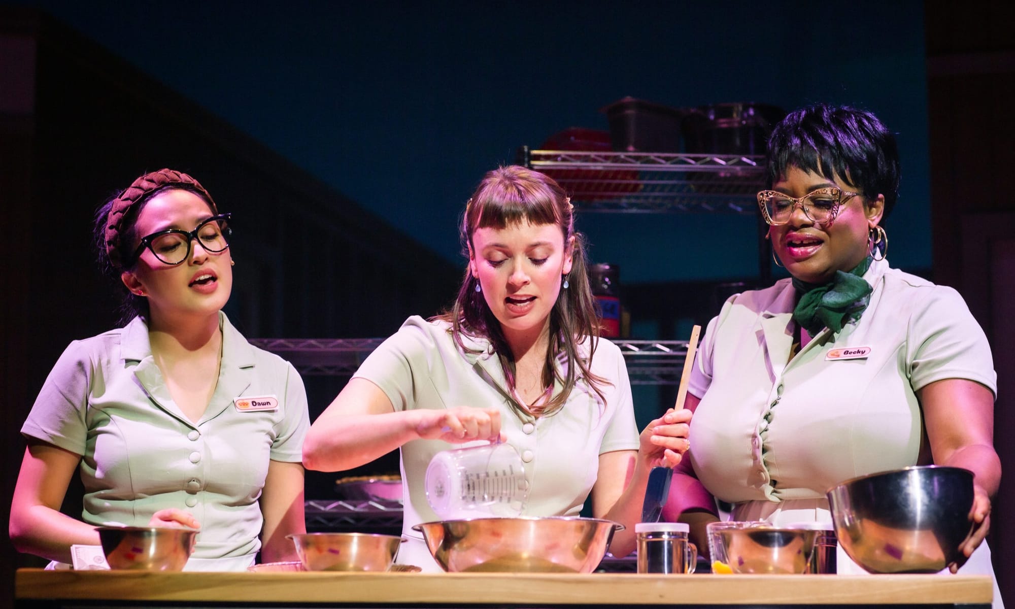 Three women wearing waitress uniforms stand in front of mixing bowls and baking material.