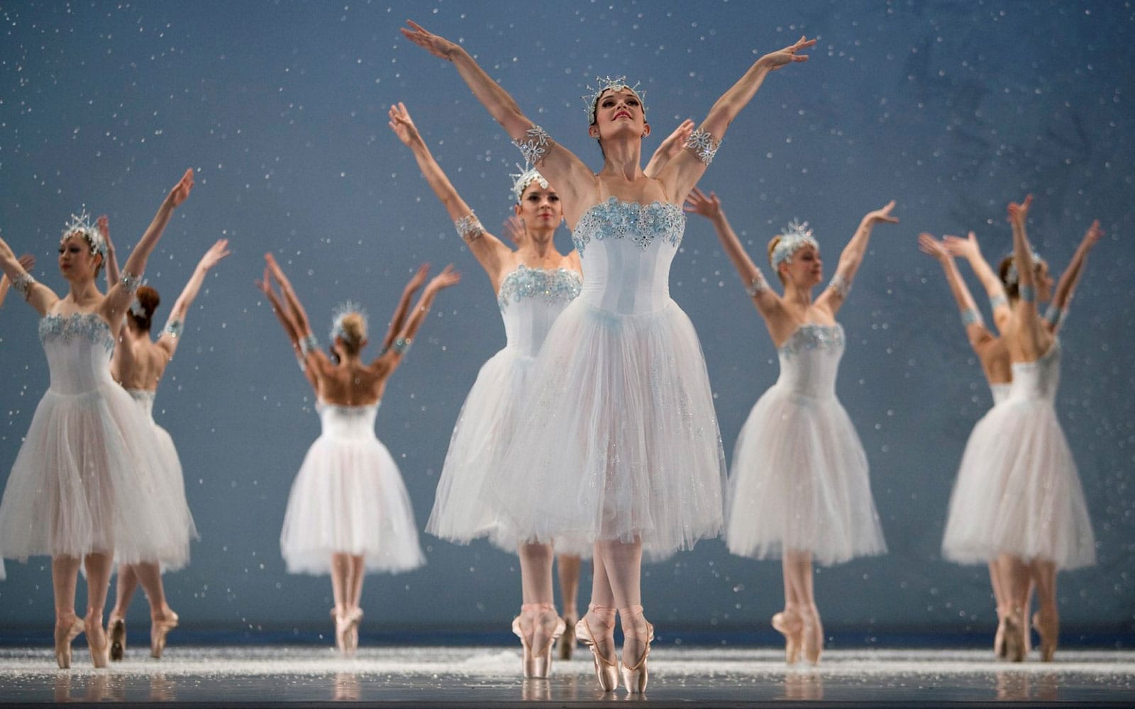 A group of ballerinas wear white gowns as snowflakes fall. They are on pointe with their arms raised. 