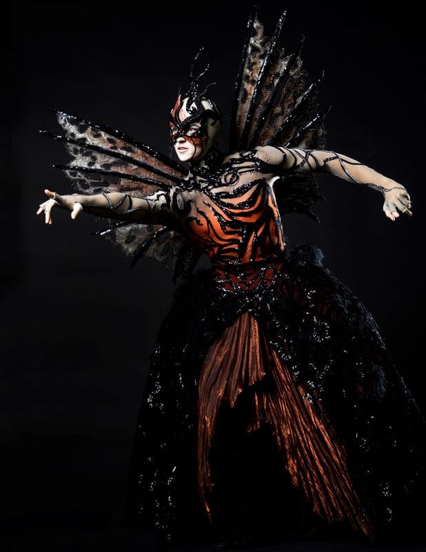 A dancer dressed in a black and orange fairy costume with a mask.