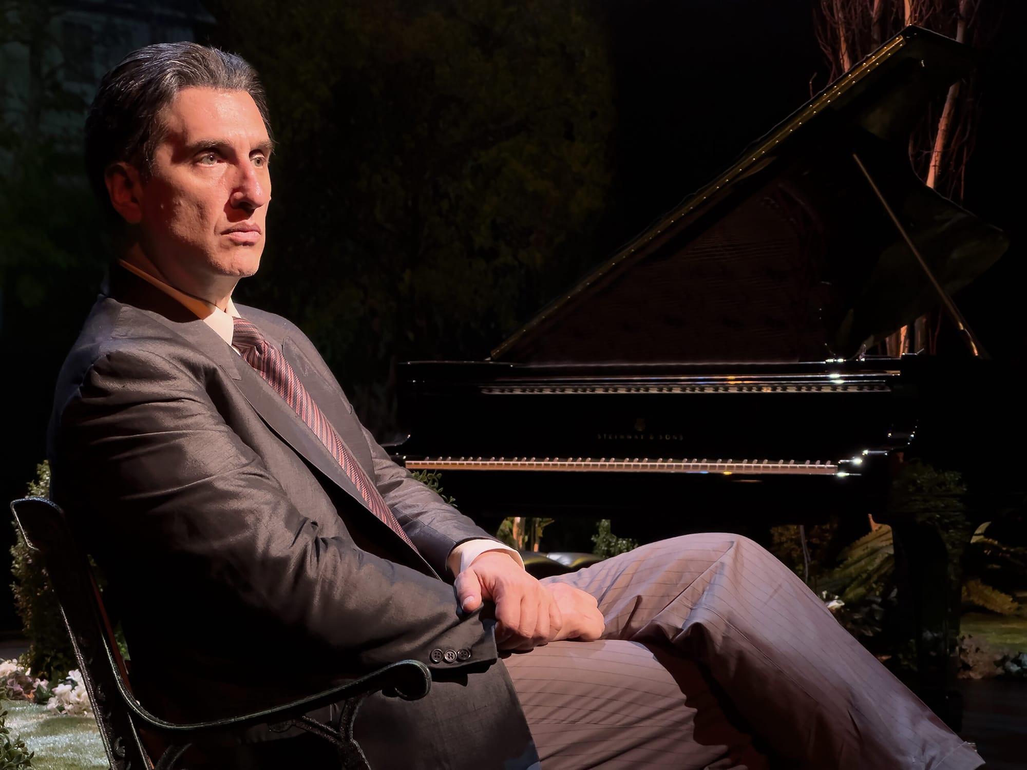 A man in a suit with pinstripe pants sits in a chair in front of a piano.