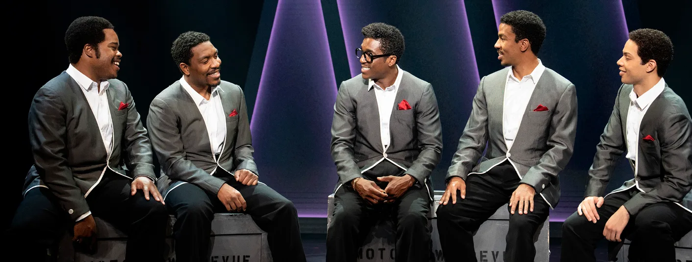 Five Black men sit talking, they're all wearing matching suits and smiling.