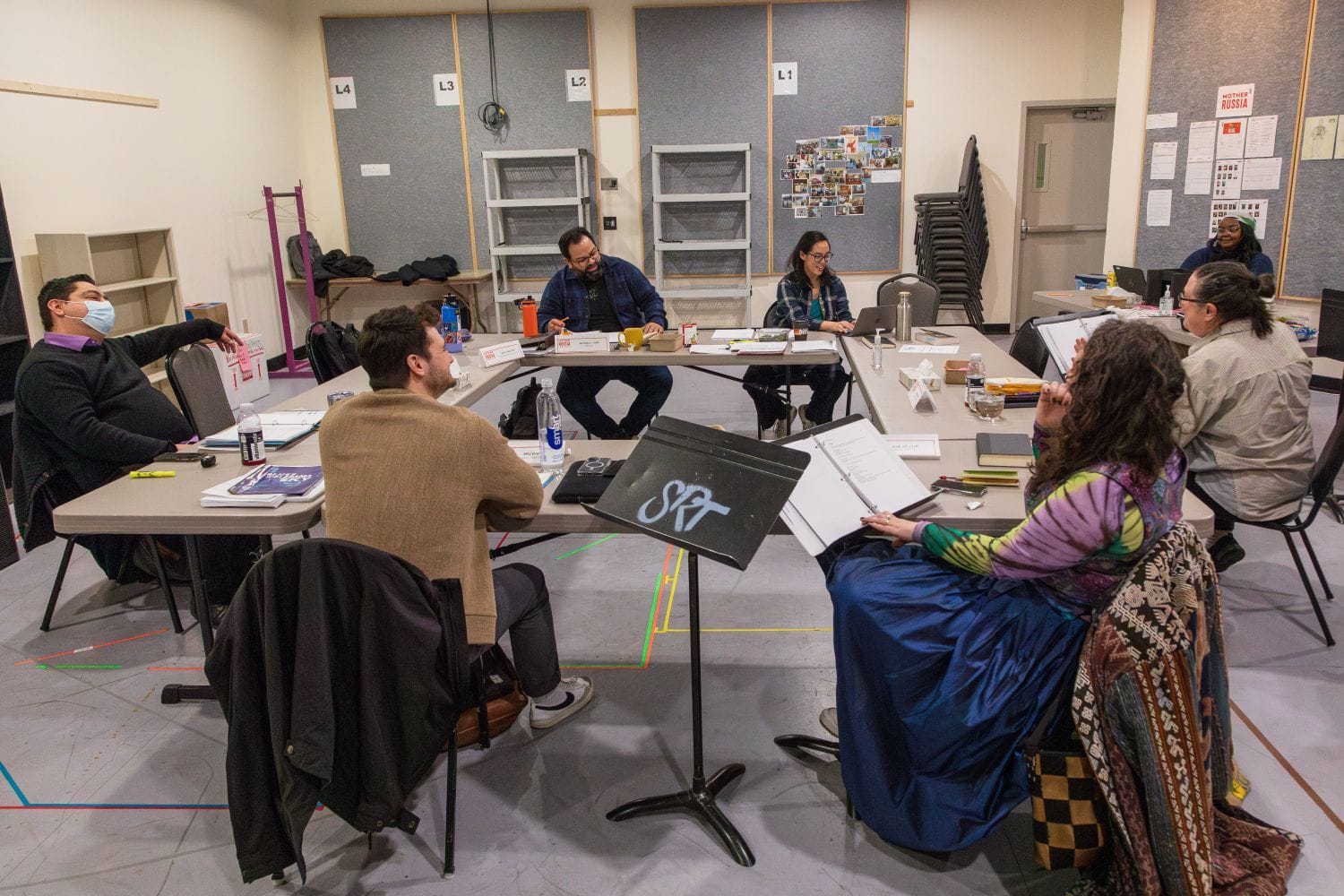 A group of people sit around a few tables reading from scripts.