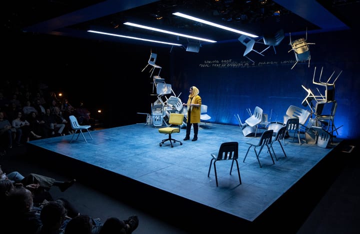 A black box theatre. A woman wearing yellow stands in front of an audience. There are chairs stacked around the stage.