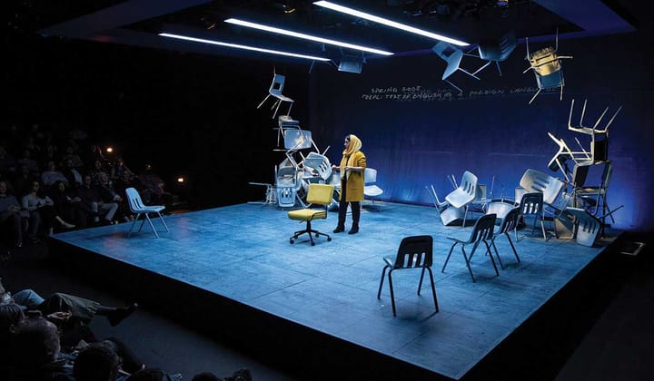 A black box theatre. A woman wearing yellow stands on stage in front of an audience. 