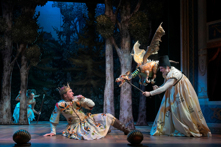 An actor holds a dragon puppet scaring a man on the ground wearing a crown. A fairy is in the background in the woods.