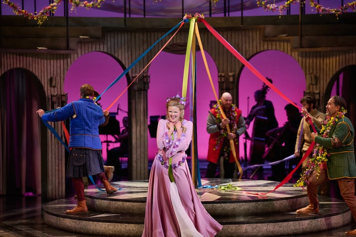 A woman in a pink medieval dress smiles joyfully in front of a may pole.