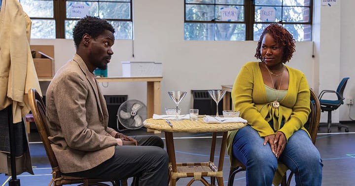 Two actors sit at a table with drinks looking at each other with skeptical faces.