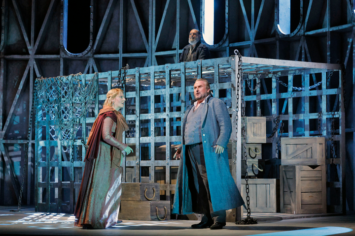 A man and woman stand talking on stage seriously in front of a jail-like storage center. 