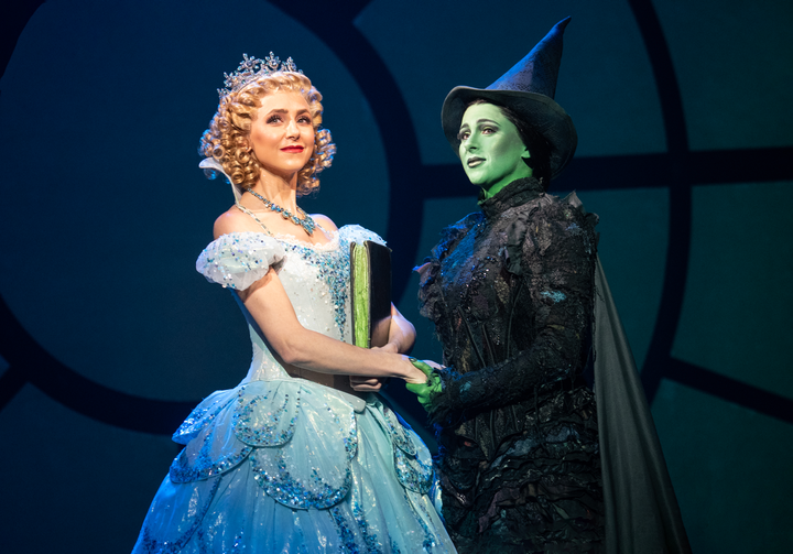 Glinda wears a blue sparkly gown and a crown and Elphaba wears a black dress and pointed hat. They are holding hands.