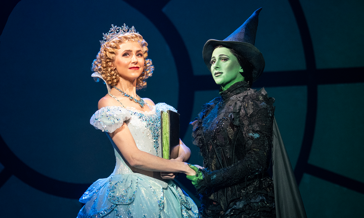 Glinda wears a blue sparkly gown and a crown and Elphaba wears a black dress and pointed hat. They are holding hands.