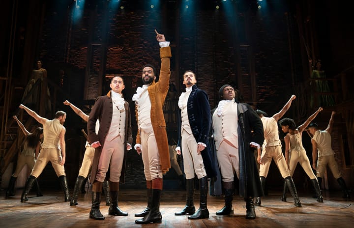 Four men wearing 18th century clothing stand on stage looking upwards. The one in front holds his arm up.