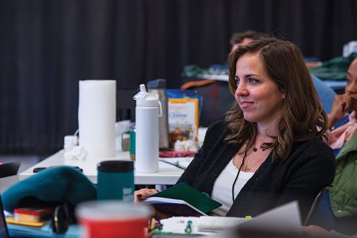 Director Kaytlin McIntyre sits smiling with a notebook in her hand watching a play rehearsal.