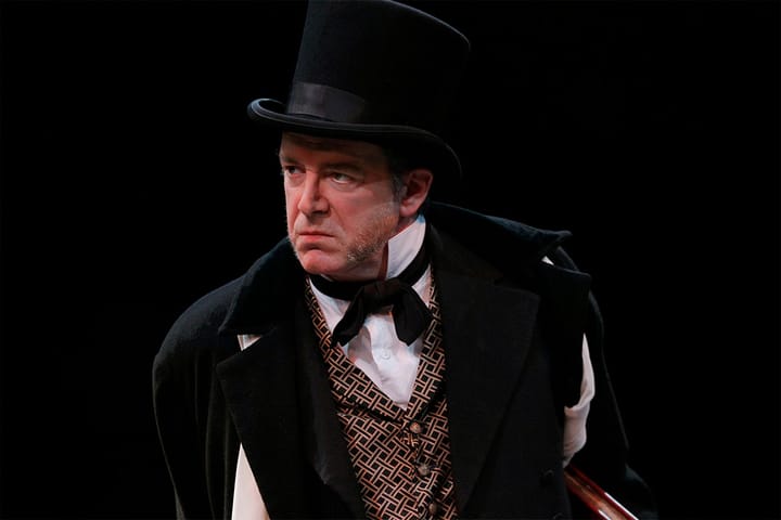 An older man dressed in 19th century clothes and a top hat frowns. 