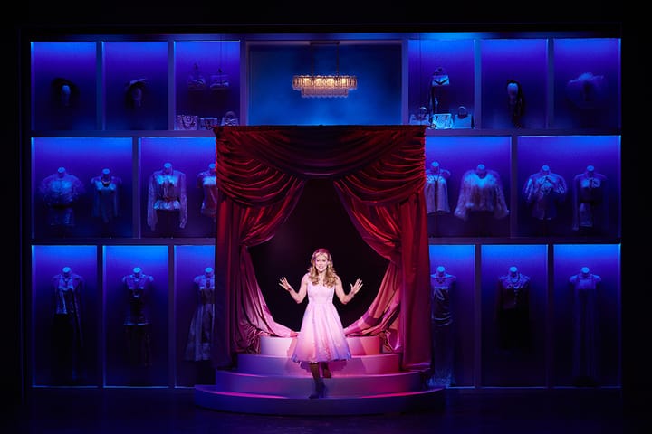 An actress wears a pink dress with a bow. She's coming down stairs. Clothes hang in the background.