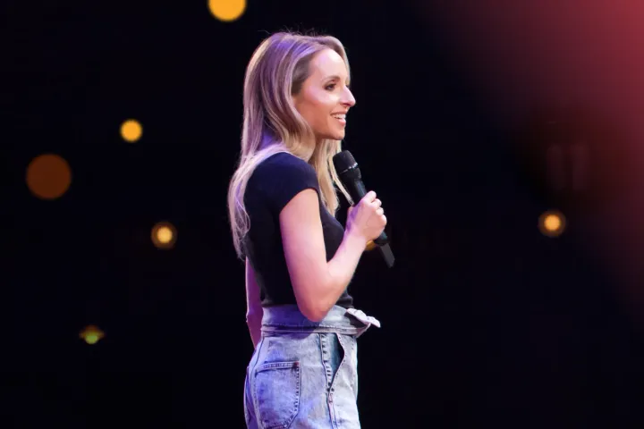 Speaker Gabby Bernstein stands holding a microphone and smiling in front of a black background.