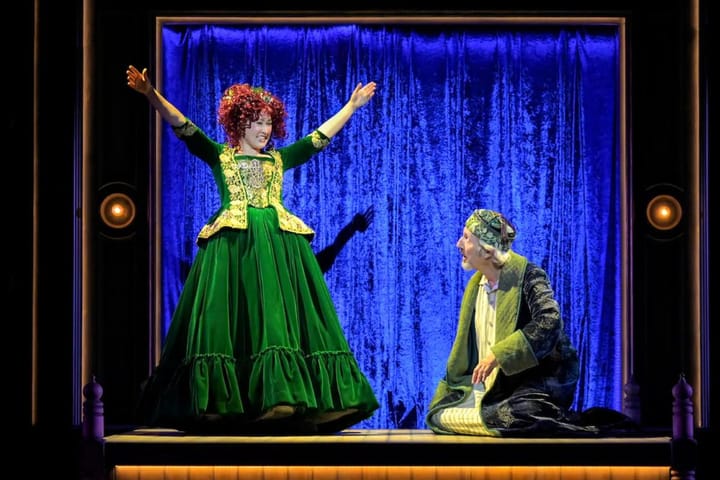 A woman in a velvet green gown smiles with her hands raised as a man in a nightgown sits on the floor looking up at her.