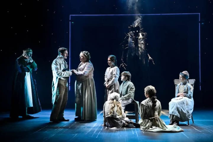 A group of actors, including adults and children, stand on a dark stage with a scary ghost puppet in the background.