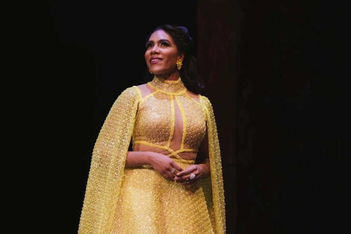 Opera singer J'Nai Bridges smiles, wearing an intricate golden dress against a black background.