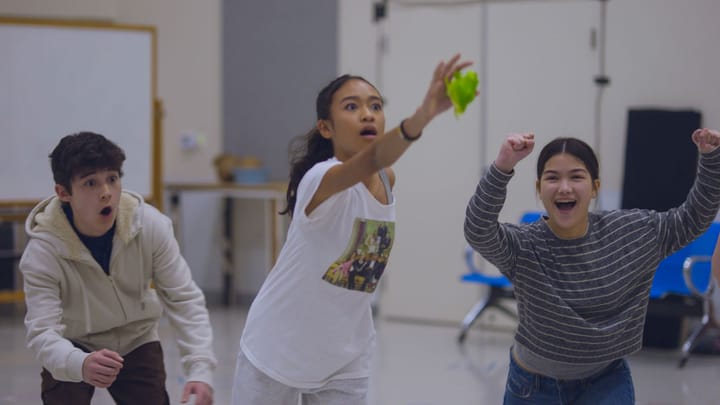 A theatre rehearsal. Three teenagers lunge forward, one looks worried and one cheers.