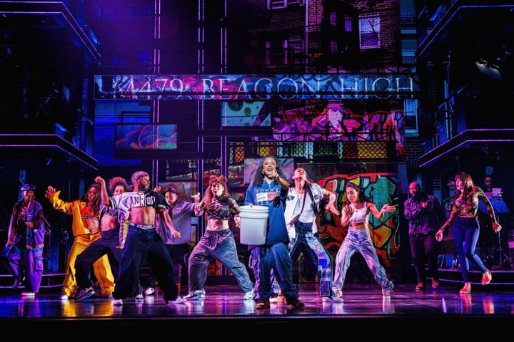 A group of dancers on stage in front of apartment buildings filled with graffiti. The woman in front holds a bucket.