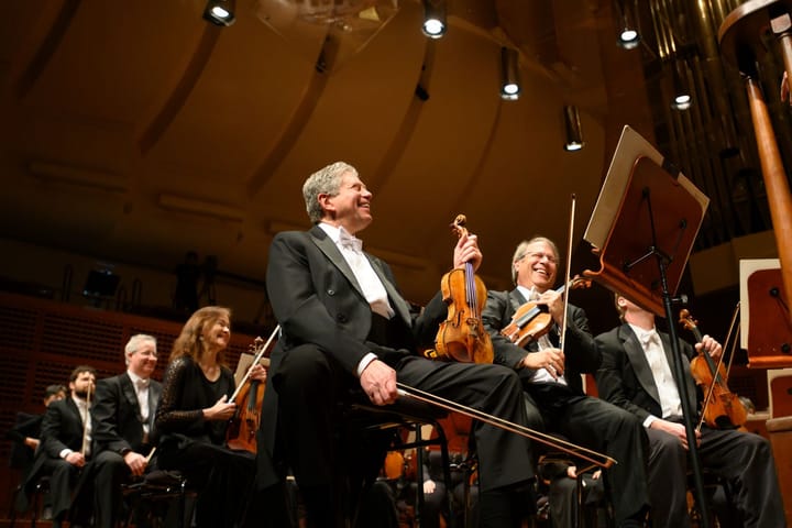 The first violin of SF symphony sits with his violin on his lap as he smiles and laughs.