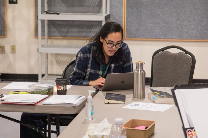 Playwright Lauren Yee sits at a table filled with scripts, looking at a laptop and working.