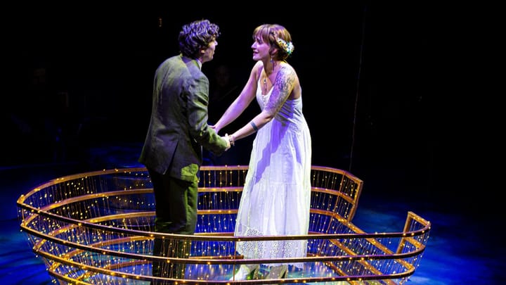 A man and woman stand holding hands. They stand surrounded by a frame with twinkle lights.